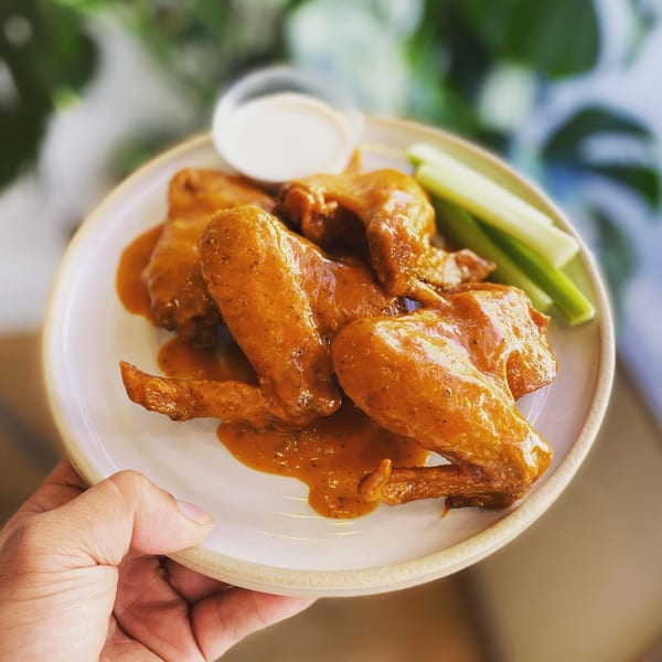 A hand holds a plate of saucy Peruvian chicken wings with celery sticks and dipping sauce.
