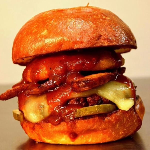 A Brazas BBQ Chicken burger with melted cheese, pickles, bacon, and red sauce on a bun.